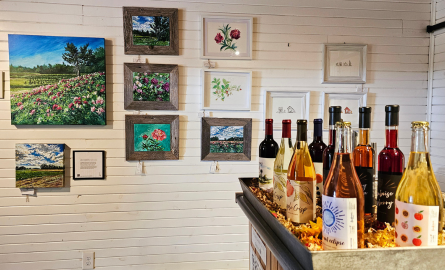 A table with a bunch of bottles of wine, on the wall is a bunch of paintings from local artists