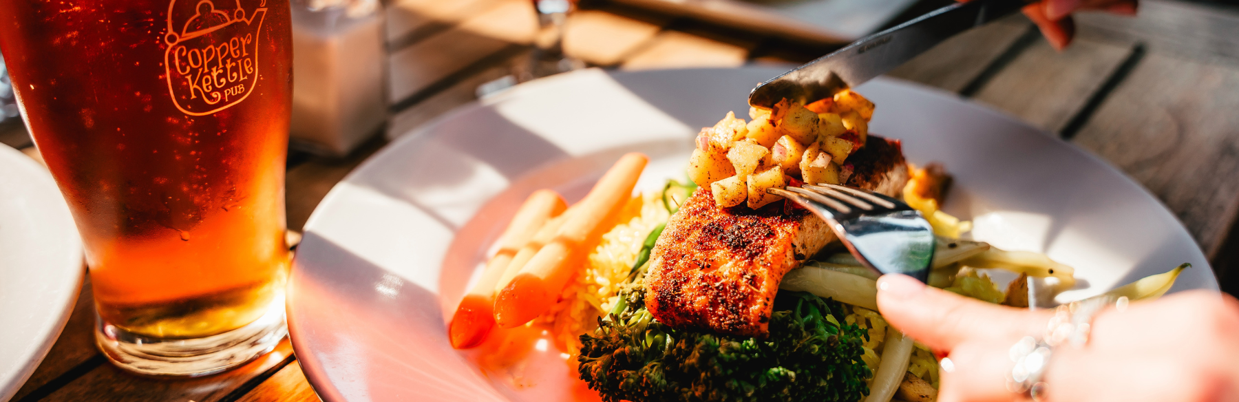 A plate of fish with someone using a fork and knife to cut it. On the left side there is a glass of beer